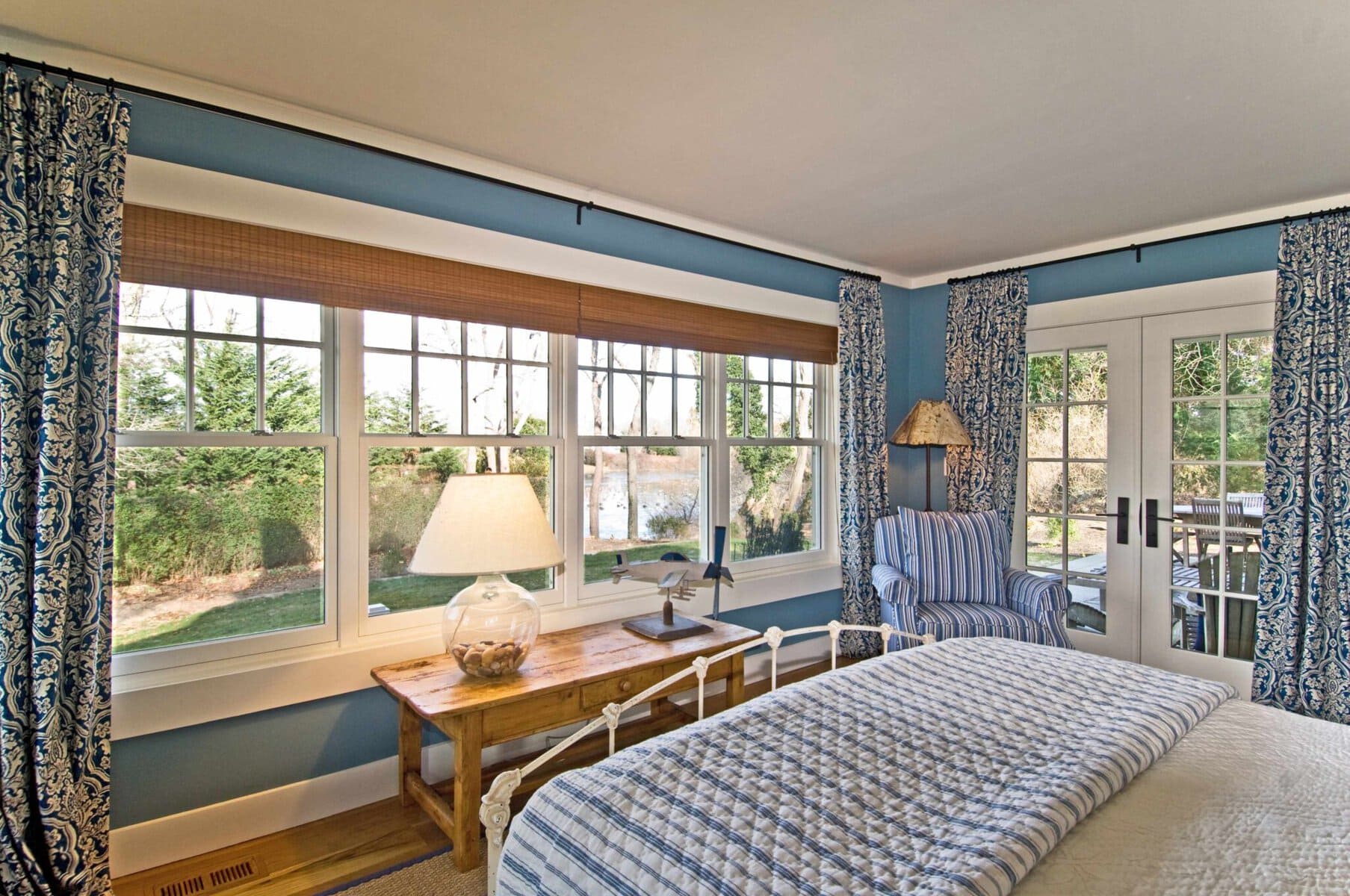 A bedroom with blue walls, patterned curtains, large windows, a wooden desk with a lamp, a striped chair, and a glass door leading outside.