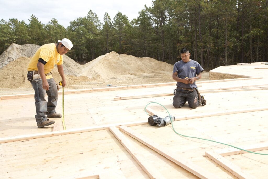Two construction workers measure and prepare wooden framing on a plywood base at an outdoor building site with sand piles in the background.