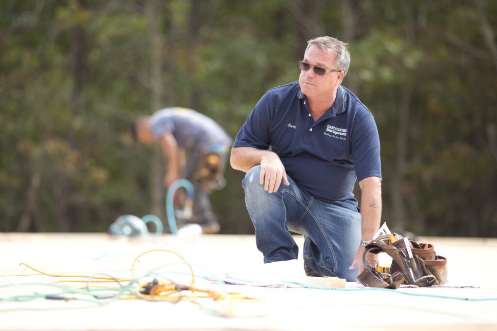 A man wearing sunglasses and a blue polo shirt kneels on a construction site, with tools and cables nearby; another worker is blurred in the background.