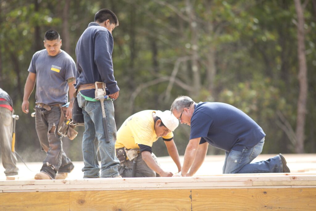 Four men work together on a wooden construction site outdoors, with tools and safety gear, surrounded by trees in the background.