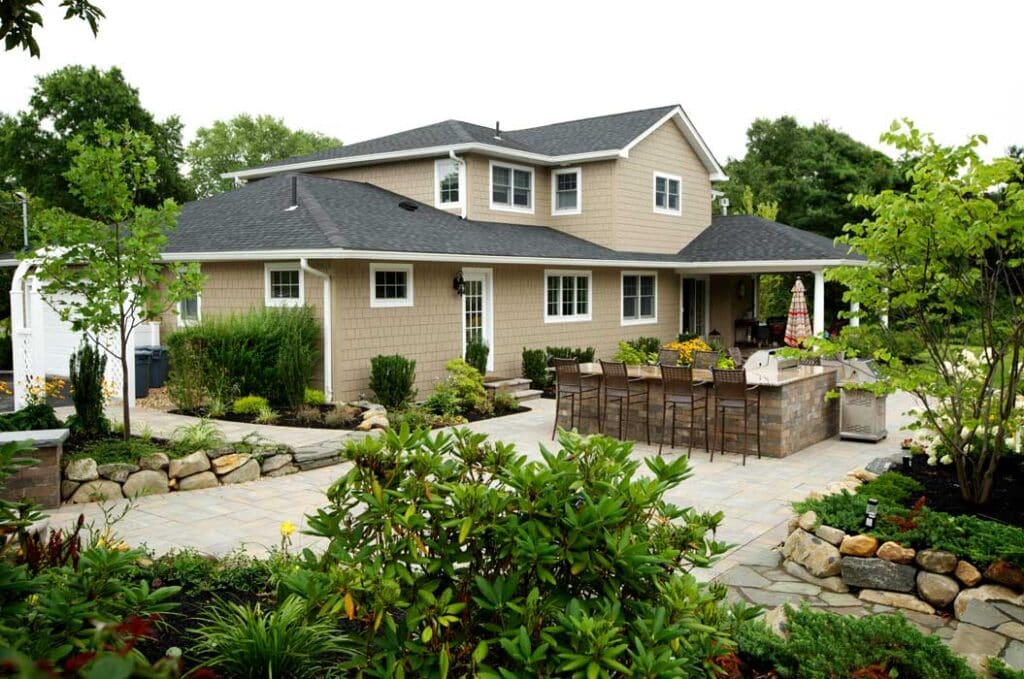 A two-story beige house with a black roof, surrounded by landscaped gardens and a stone patio with outdoor seating and a grill area.