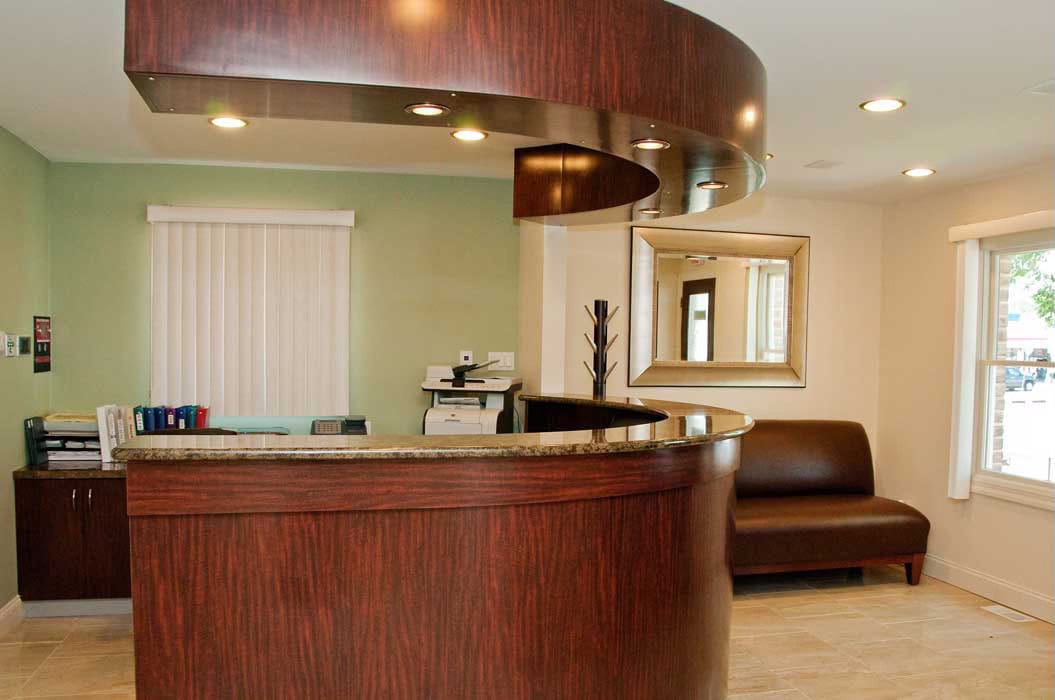 Reception area with a curved wooden desk, office equipment, a brown bench, a large wall mirror, and windows letting in natural light.