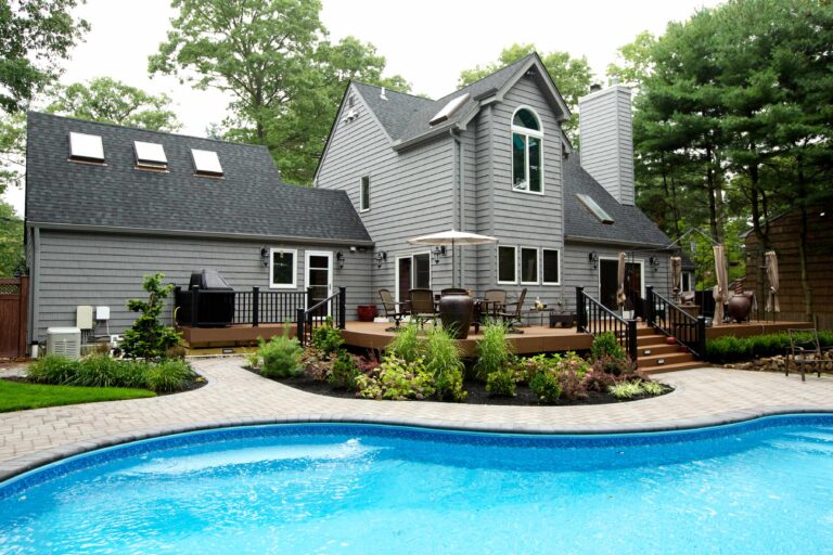 A modern gray house with a wooden deck, outdoor seating, grill, landscaped garden, and an in-ground swimming pool in the backyard.