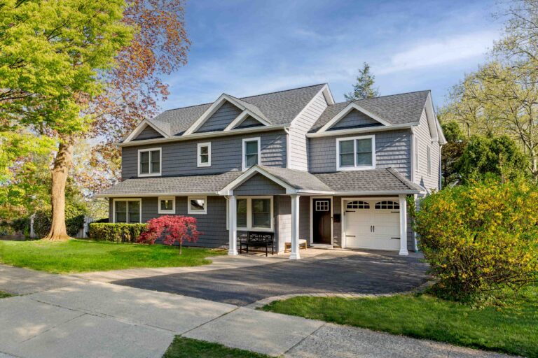 Two-story gray suburban house with white trim, attached garage, and front porch, surrounded by trees and shrubs on a sunny day.
