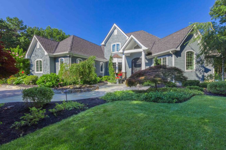 A large gray house with gabled roofs, surrounded by green landscaping, trees, and a manicured lawn under a clear blue sky.