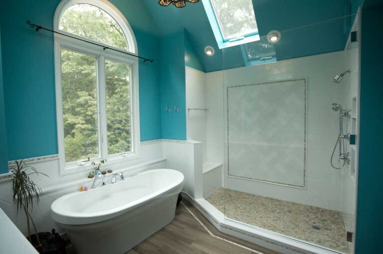 Modern bathroom with turquoise walls, a freestanding white bathtub by a window, a large walk-in shower with pebble floor, glass divider, and a skylight above.