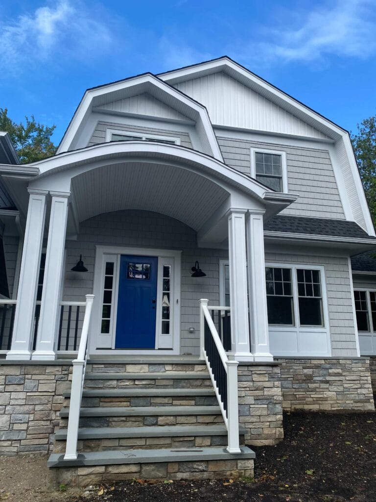 Two-story house with gray siding, white trim, stone foundation, front porch with columns, and blue front door under a covered entry. Steps lead up to the entrance.