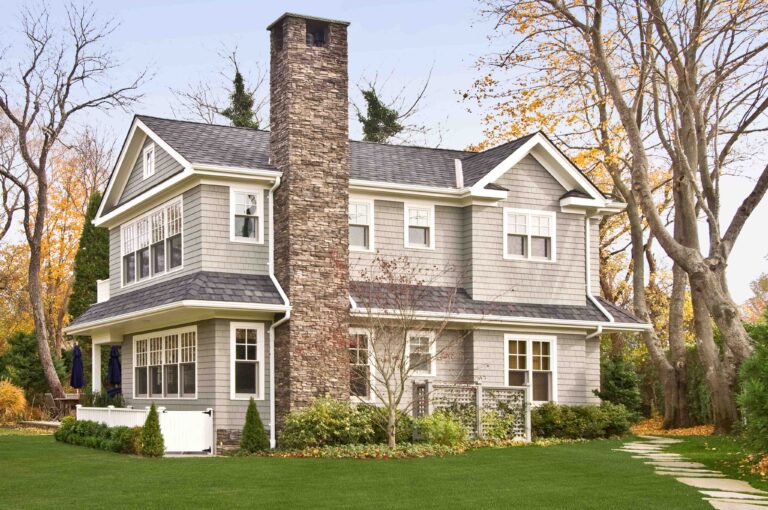 Two-story gray house with white trim, stone chimney, large windows, and a well-maintained lawn, surrounded by trees with autumn leaves.