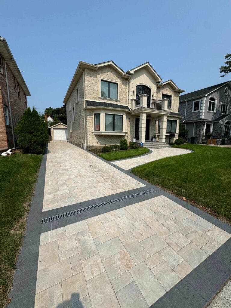 Two-story beige brick house with a double garage, paved driveway, front yard with green grass, and neighboring homes on both sides.