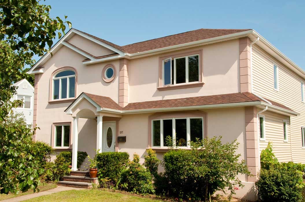 Two-story beige suburban house with a brown shingle roof, white trim, front porch, and surrounded by green bushes and lawn on a sunny day.