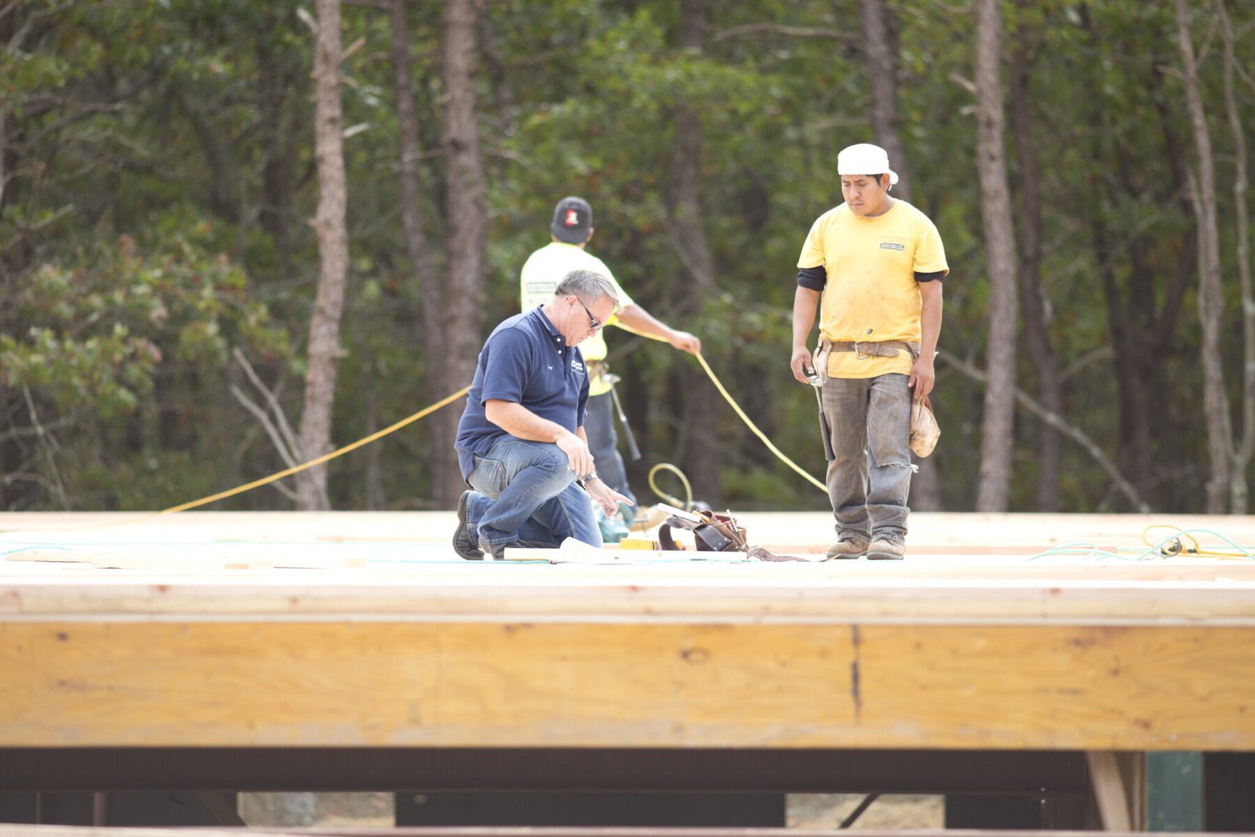 Three men work on a wooden construction site; one kneels inspecting tools while two others stand, with trees visible in the background.