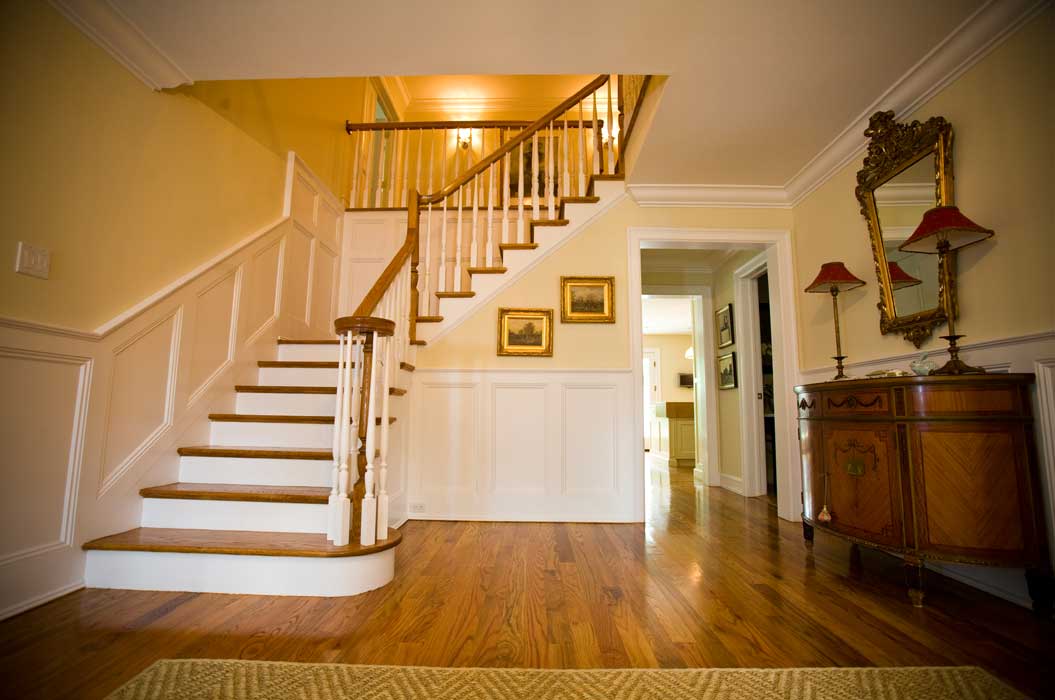 A wooden staircase with white trim in a hallway featuring hardwood floors, framed artwork, a mirror, and a wooden sideboard with two red lamps.
