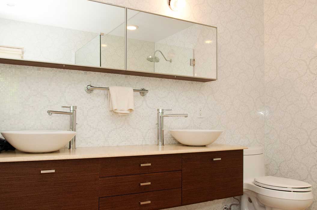 Modern bathroom with double sinks, a wooden vanity, wall-mounted mirrors, a towel rack with a white towel, and a toilet. Subtle patterned wallpaper covers the walls.