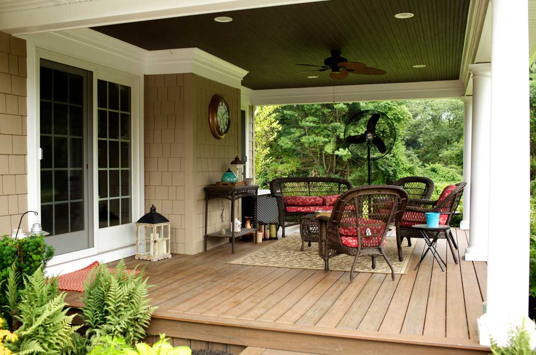 Covered wooden porch with wicker furniture, red cushions, a small table, ceiling fan, plants, and a view of green trees in the background.
