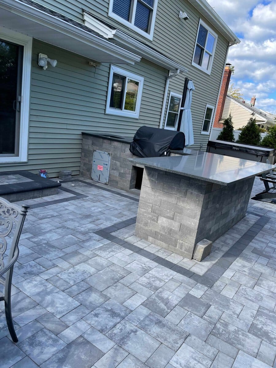 A backyard patio with gray stone pavers, an outdoor kitchen island, a grill, patio furniture, and a two-story house in the background.